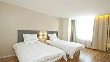 A hotel room with two beds, a wooden floor, a large window with curtains, and a small table.
