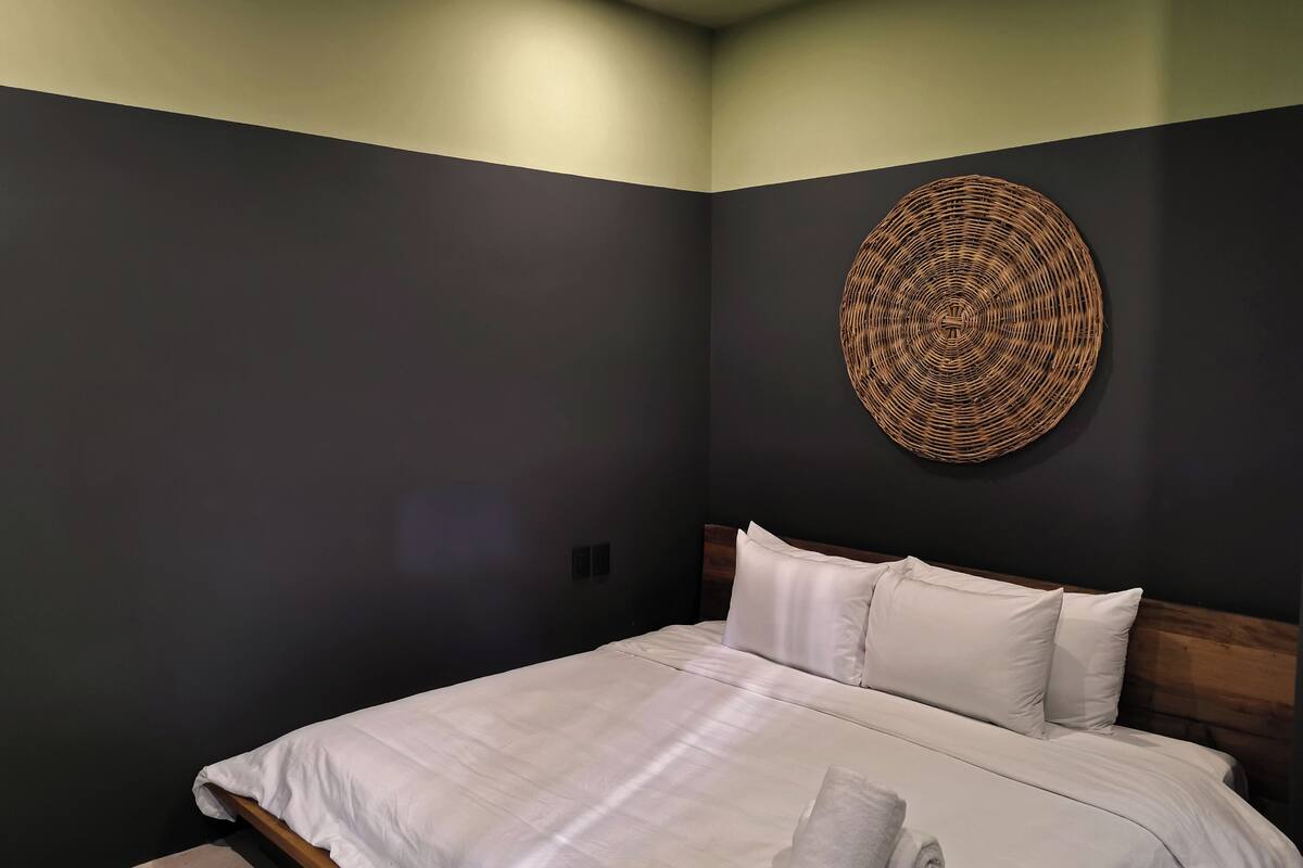 A neatly made bed with white linens and pillows, a rolled white towel, and a woven wall art piece.