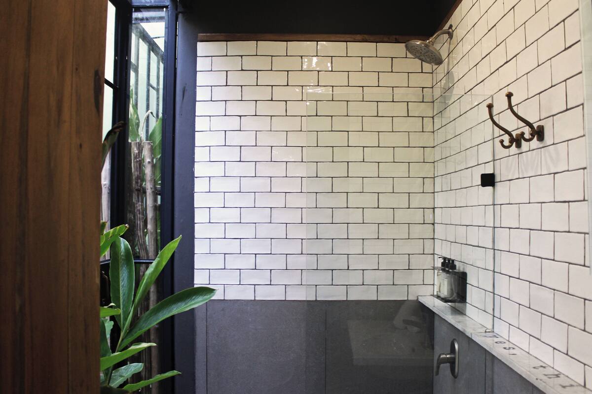 A bathroom with white and gray tiles, a shower, a plant, and a small shelf with toiletries.