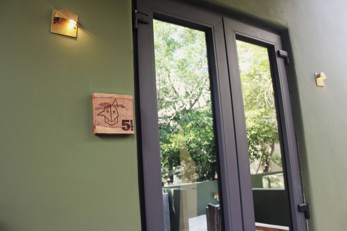 A door with a number sign indicating room 5, a light fixture above it, and a view of greenery outside.