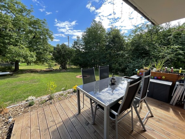 Um deck de madeira com mesa e cadeiras, com vista para uma área gramada com árvores e uma mesa de piquenique ao fundo.