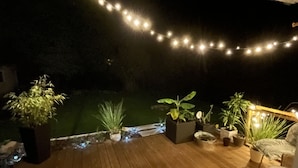Una terraza de madera con luces colgantes, plantas en macetas y mobiliario exterior.