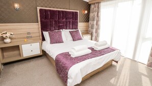 Ein modernes Hotelzimmer mit einem großen Bett, einem Holzkopfende, einem Nachttisch, einem kleinen Tisch und einem großen Fenster mit Vorhängen.