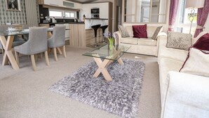Ein modernes Wohnzimmer mit einem Glastisch, grauen Sesseln, einem beigefarbenen Sofa und einer Küche im Hintergrund.