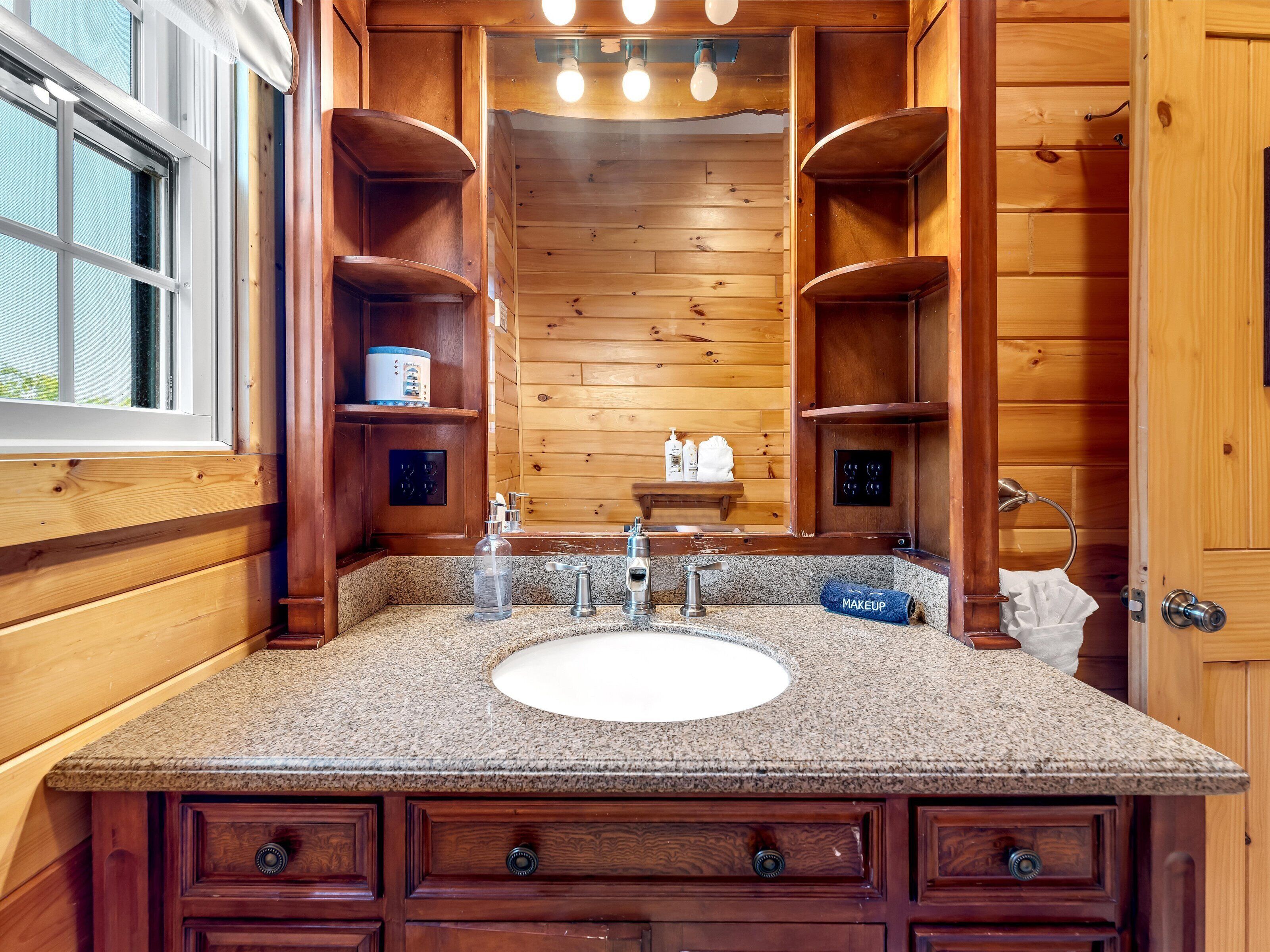 A wooden bathroom with a granite countertop, a sink, and a mirror. There are shelves with toiletries and a towel rack.