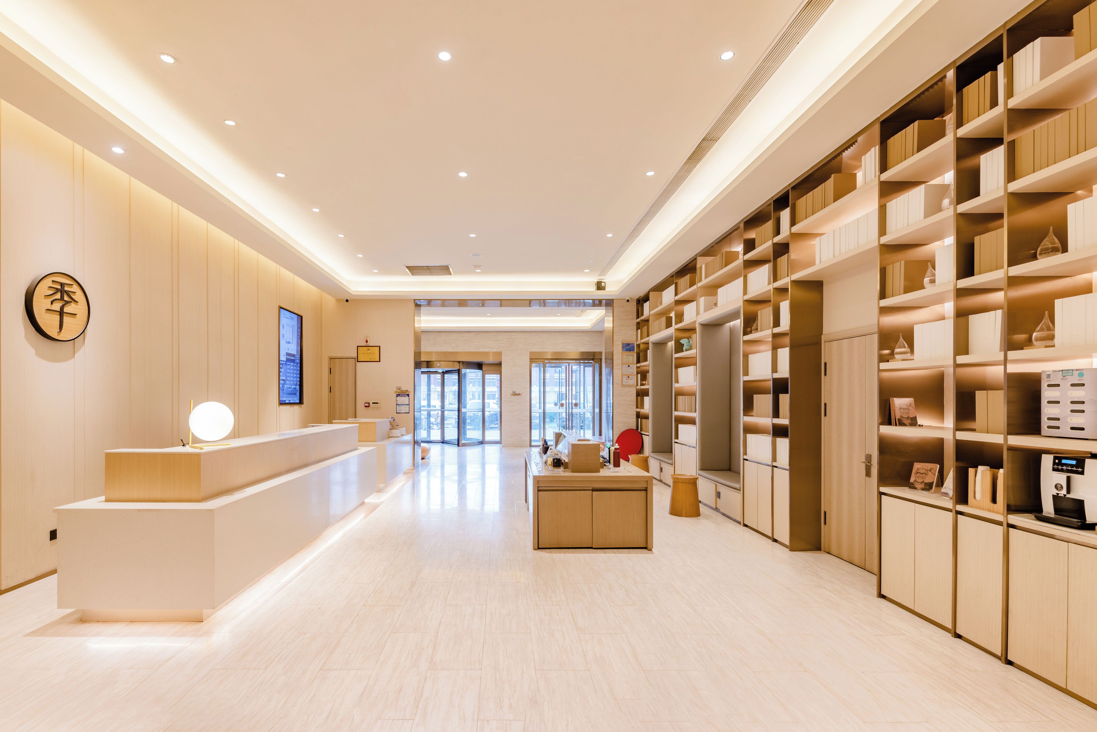 A modern hotel lobby with a reception desk, a digital screen, and a large bookshelf.