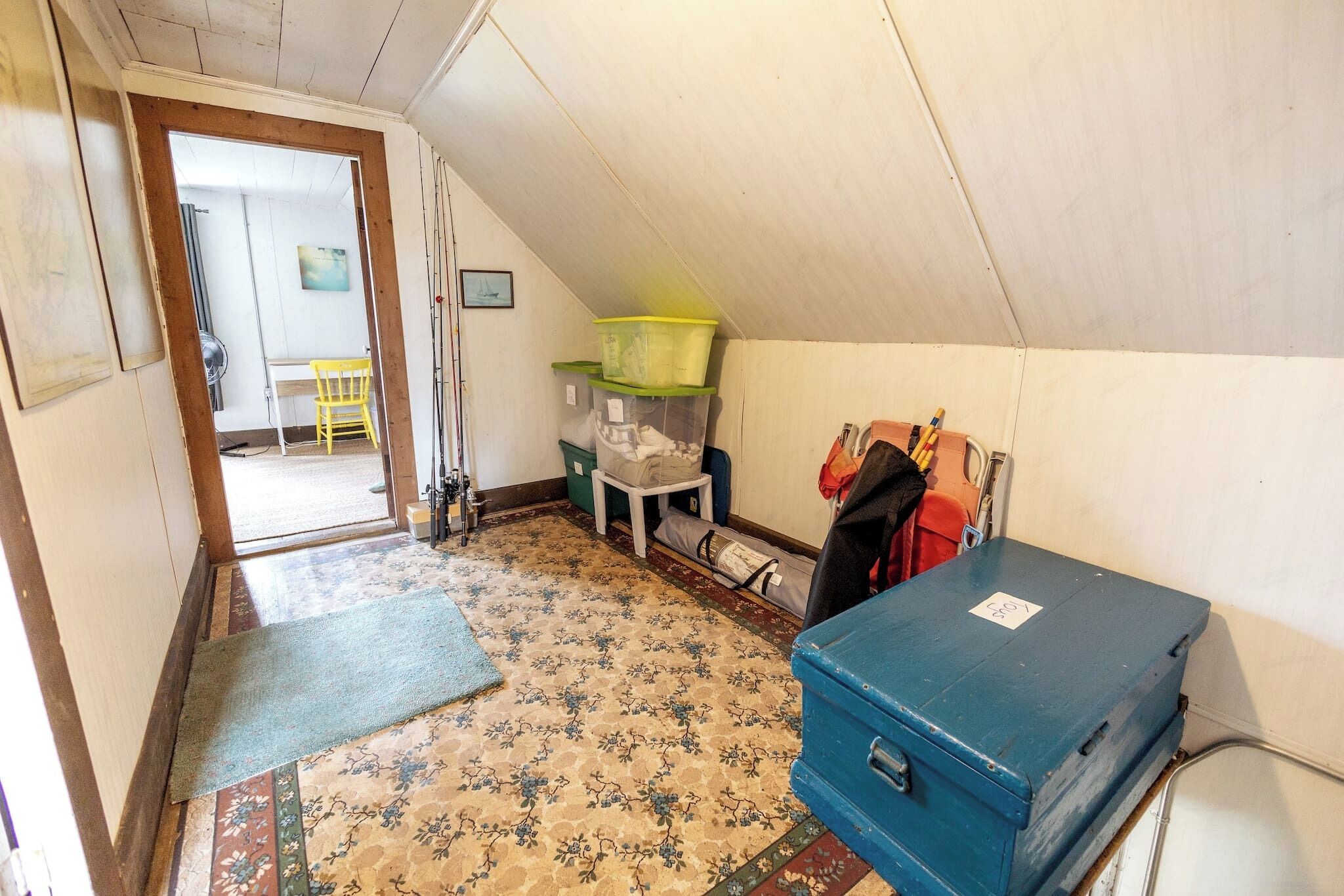 A small, narrow room with a sloped ceiling, a patterned carpet, a blue storage box, and a chair.