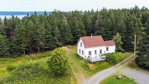 Une maison blanche avec un toit rouge, entourée d’une forêt dense, un chemin de gravier menant à la maison, et une étendue d’eau en arrière-plan.