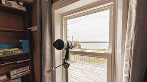 Une pièce avec une fenêtre donnant sur un plan d’eau, une bibliothèque contenant divers objets et un télescope installé près de la fenêtre.