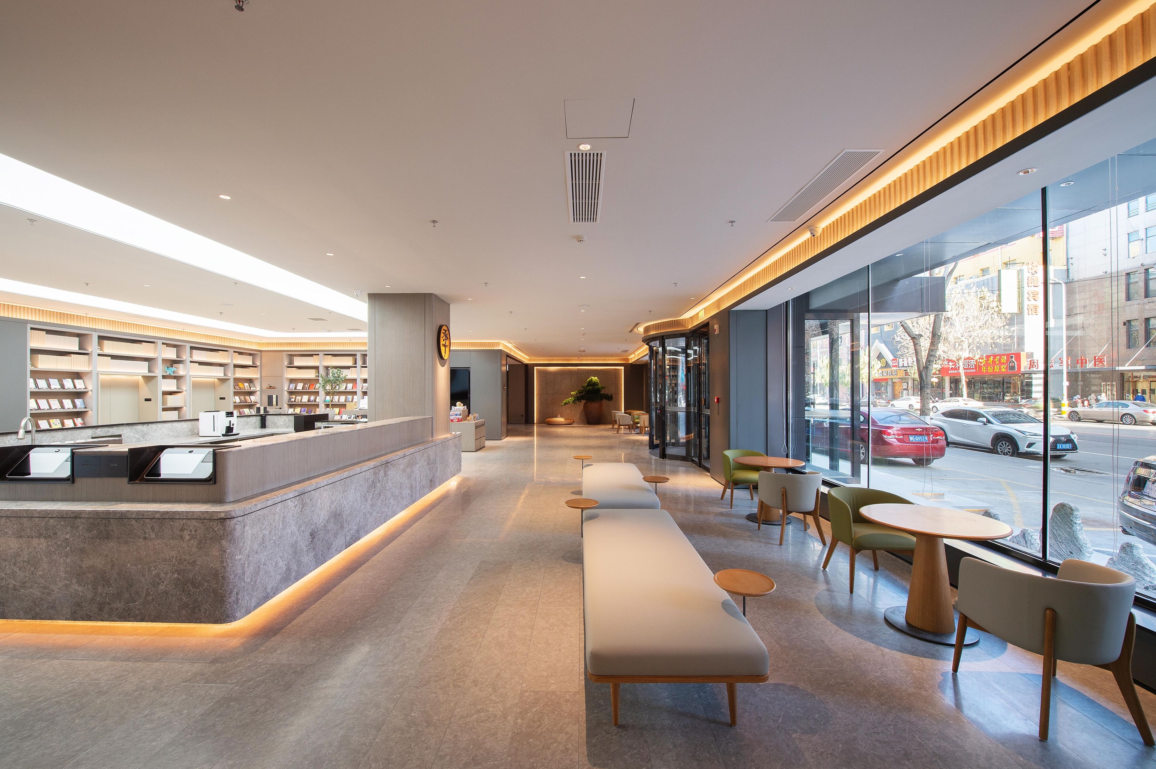 A modern hotel lobby with a reception desk, seating area, and large windows offering a view of the street.