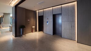 Um saguão moderno de hotel, com balcão de recepção, uma planta pequena e um elevador.