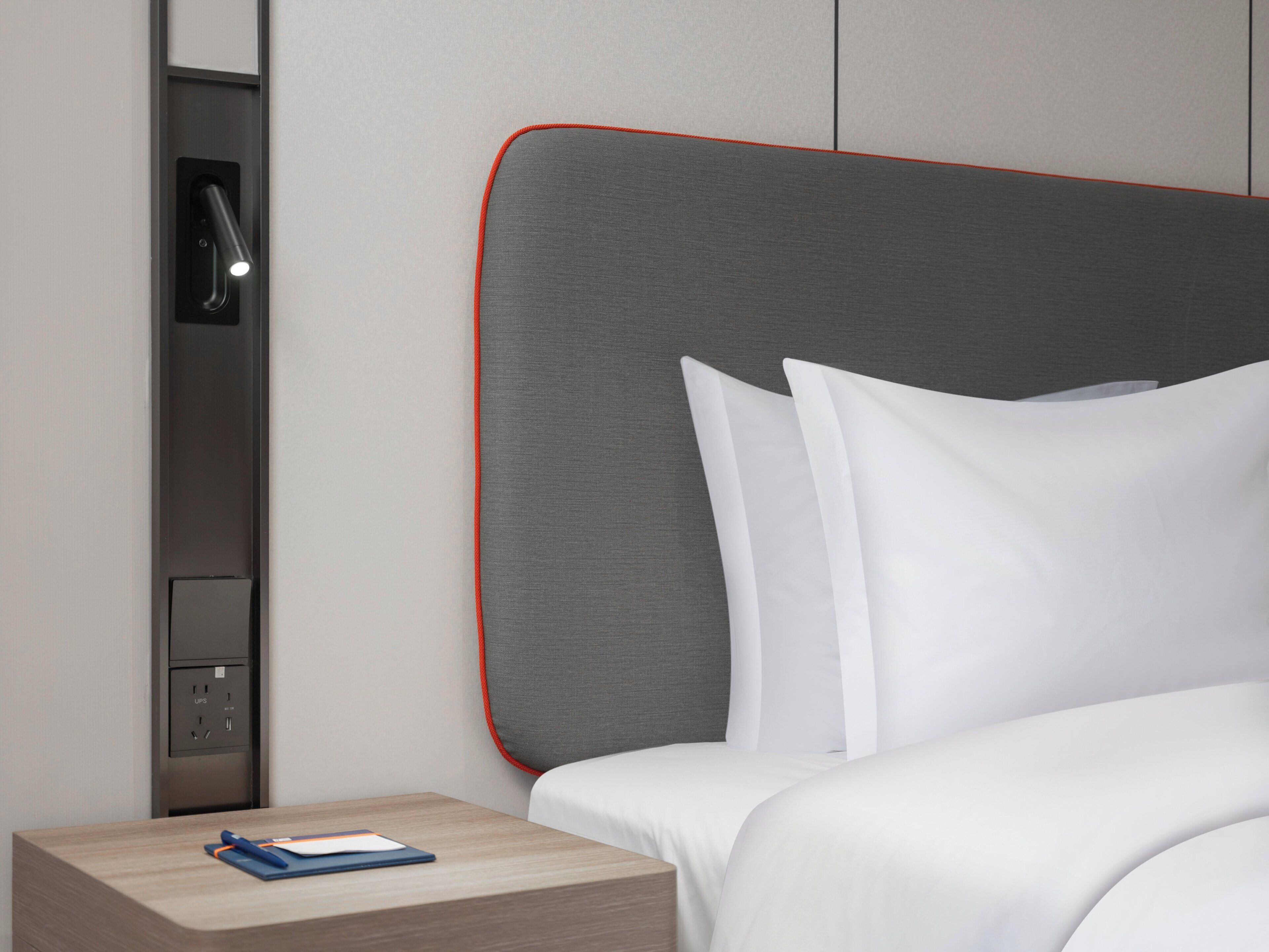 Una habitación de hotel moderna con una cama bien hecha, una mesita de noche con una lámpara y un pequeño escritorio con una computadora portátil.