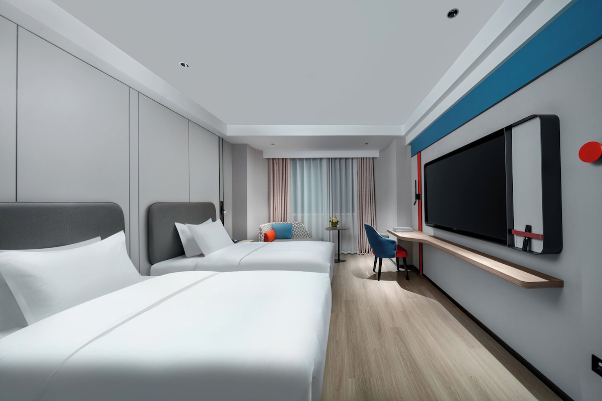 A modern hotel room with two beds, a desk, a chair, and a large TV on the wall.