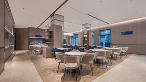 Una hall d'albergo moderna con tavoli e sedie rotonde, una scala centrale e vista sulla città attraverso ampie finestre.