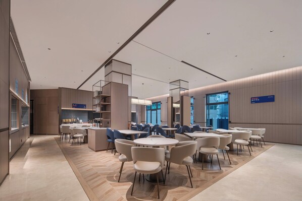 Eine moderne Hotellobby mit runden Tischen und Stühlen, einer zentralen Treppe und Blick auf die Stadt durch große Fenster.