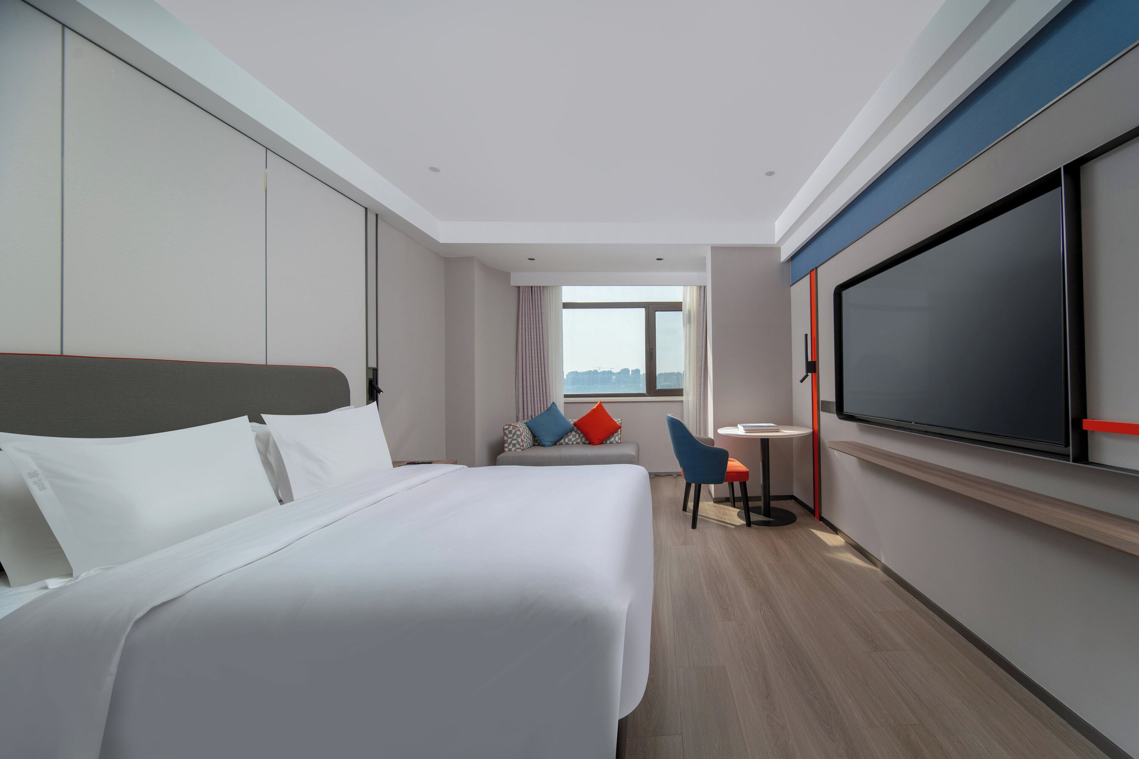 A modern hotel room with a large bed, a TV, a desk, and a view of the city.