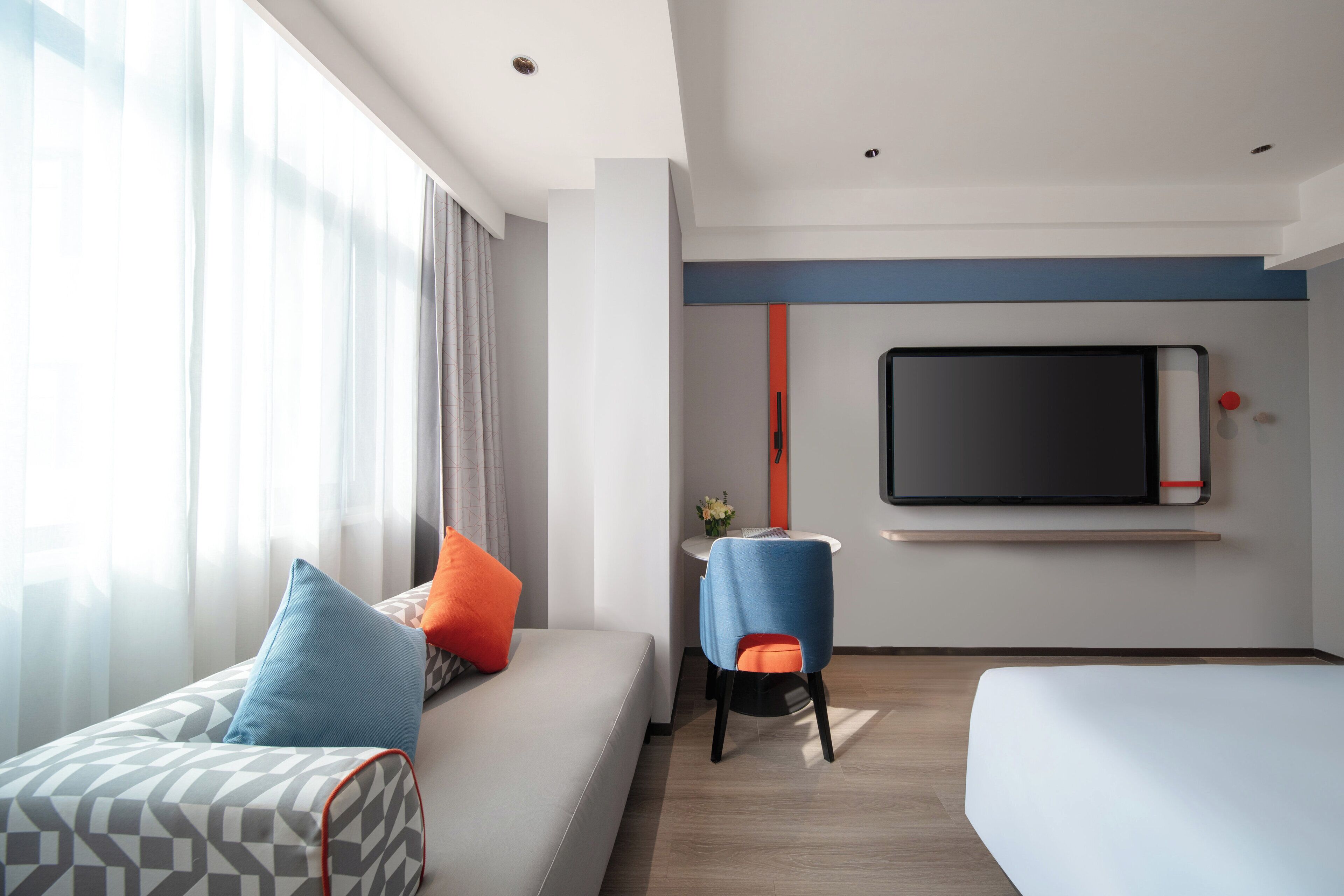 A modern hotel room with a sofa, a chair, a TV mounted on the wall, and a small table with a vase of flowers.