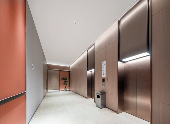 Un ascenseur moderne doté d’un écran numérique, d’une poubelle et d’une plante en pot, situé dans un couloir dont le sol est carrelé et les murs sont en bois.