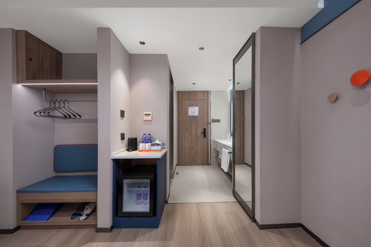 A modern hotel room with a built-in bench, a small fridge, a mirror, and a wooden wardrobe.