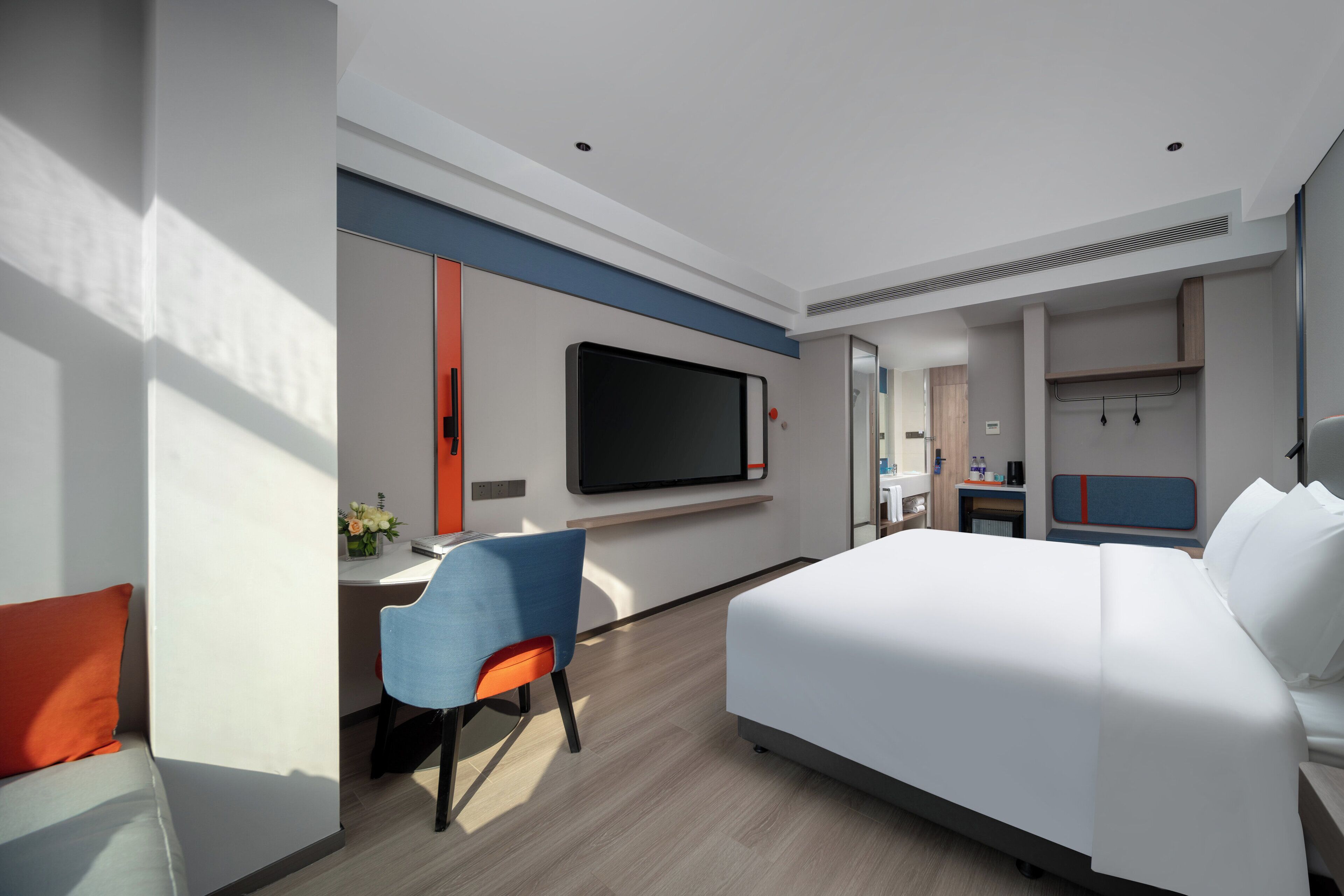 Une chambre d’hôtel moderne dotée d’un grand lit, d’un bureau avec une chaise, d’un téléviseur à écran plat fixé au mur et d’un petit coin cuisine.