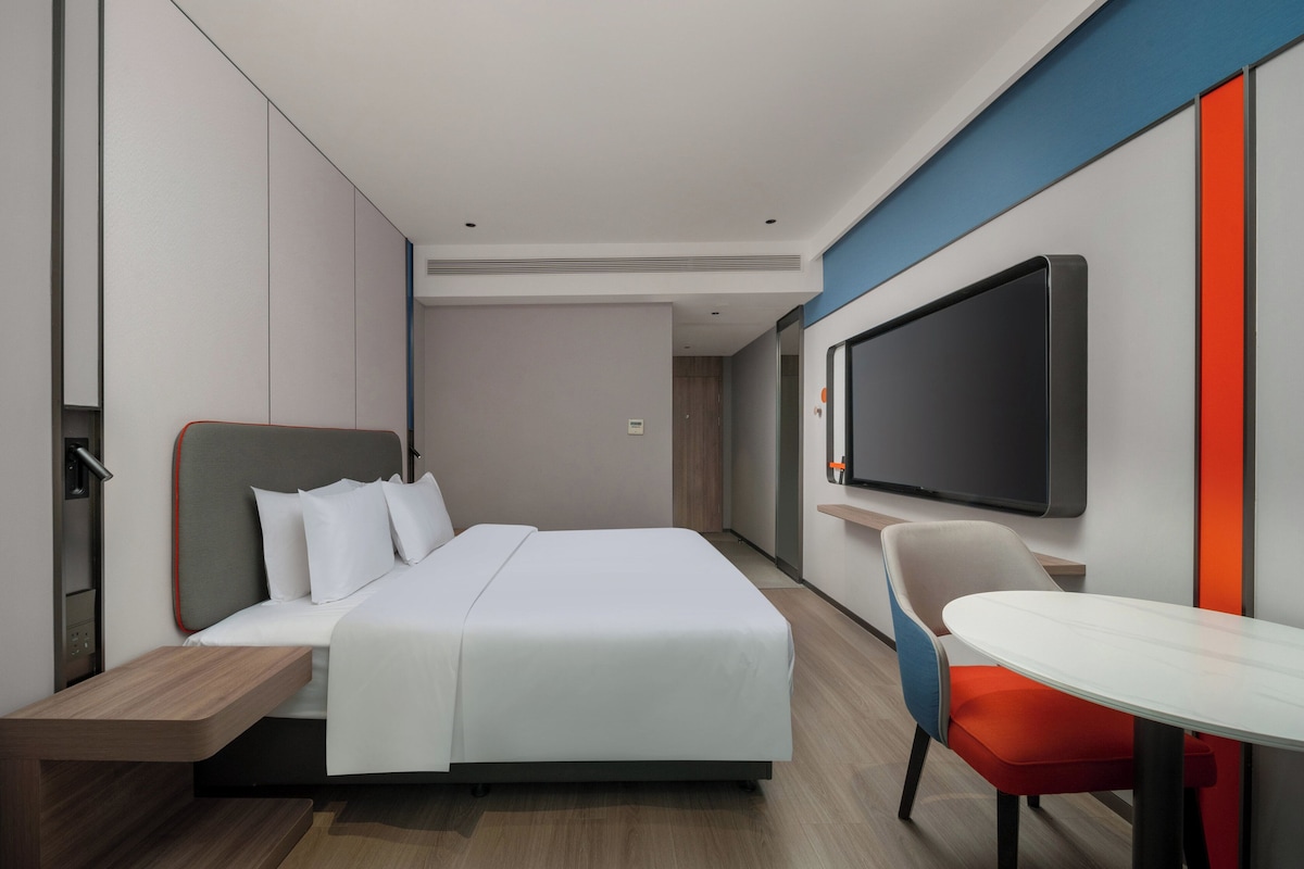 A modern hotel room with a bed, a TV mounted on the wall, a small table, and a chair.