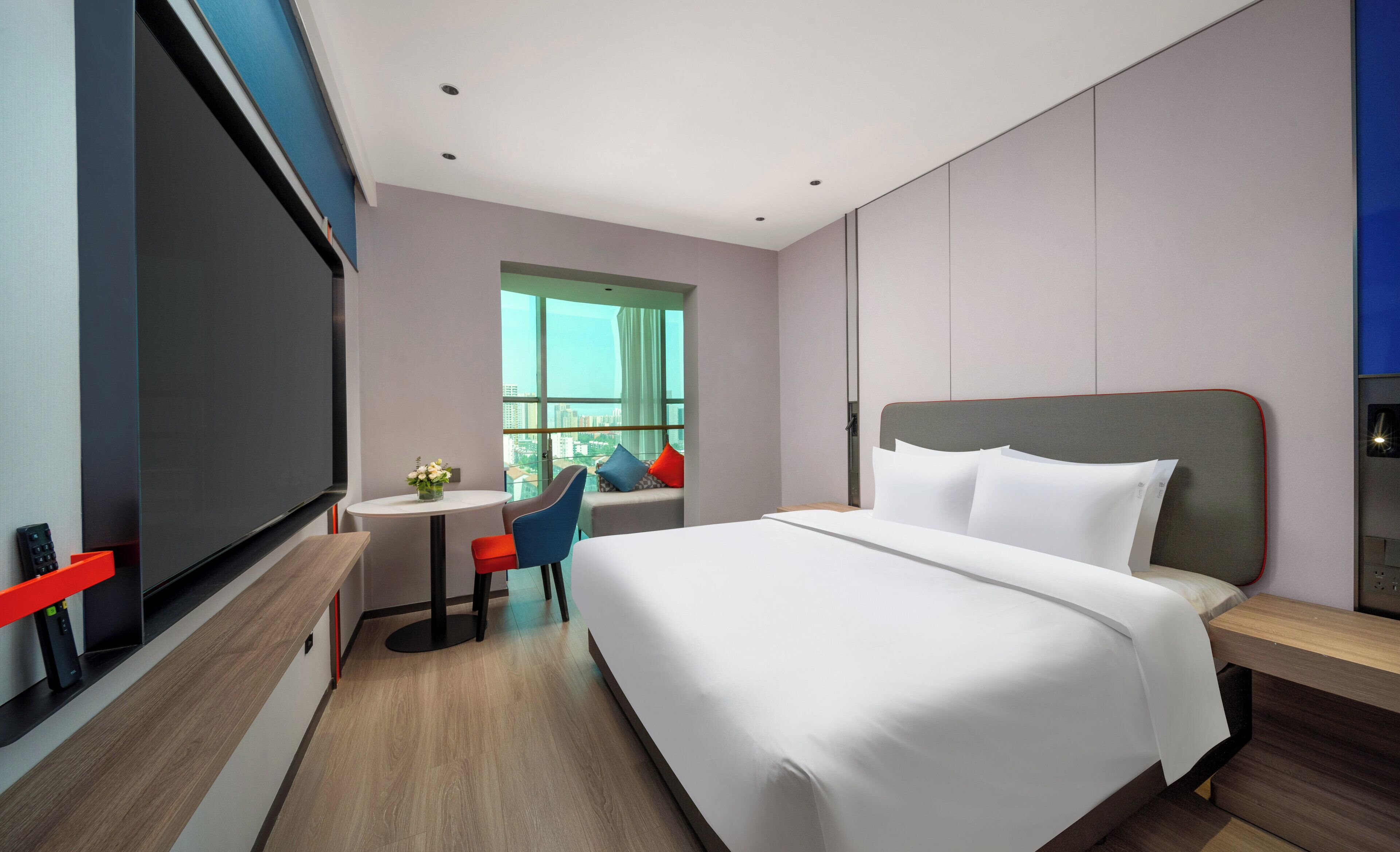 A modern hotel room with a large bed, a desk with a chair, a television, and a window with a city view.
