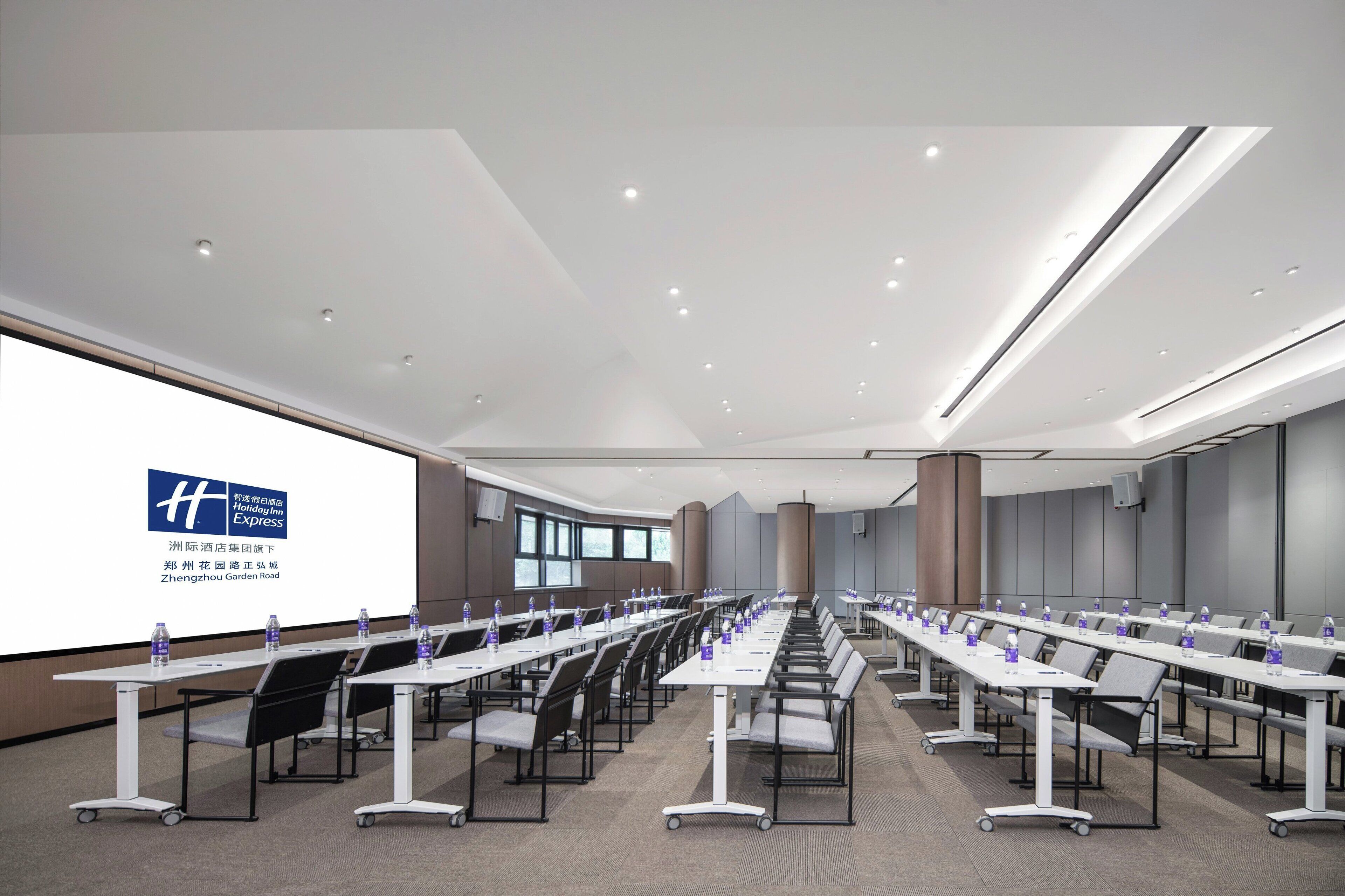 A conference room with multiple tables and chairs, a large screen displaying a logo, and a modern ceiling design.