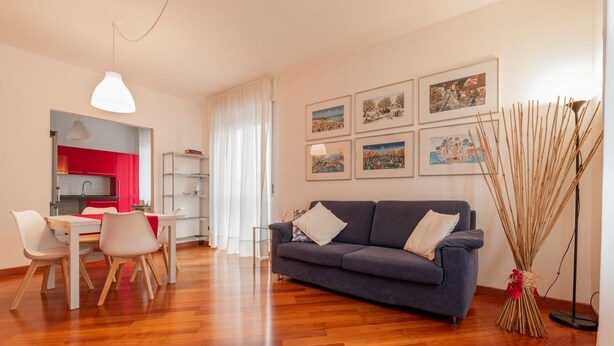 A modern living room with a grey sofa, white chairs, a dining table, and a kitchen in the background.