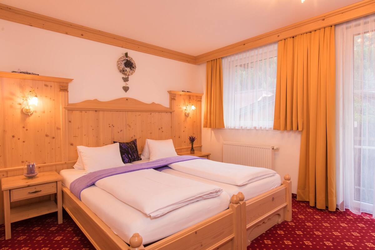 Una cama doble con cabecera de madera, dos mesitas de noche, una ventana con cortinas amarillas y una lámpara fijada a la pared.