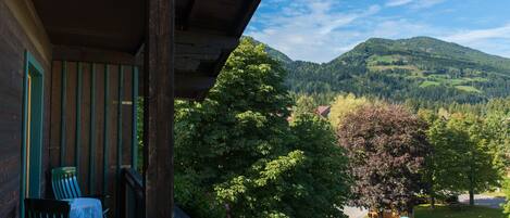 A wooden balcony with a table and chairs, overlooking a lush green landscape with trees and a mountain in the background.