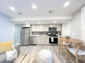 Une cuisine moderne équipée d’appareils électroménagers en acier inoxydable, un coin repas avec une table et des chaises en bois, et un espace salon confortable avec un canapé et un pouf.