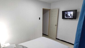 Quarto com cama, travesseiros, televisão na parede e uma porta ao fundo.