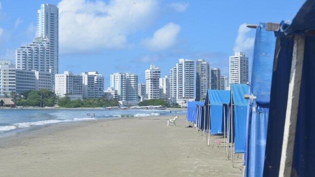 Uma praia com uma fileira de cabanas azuis e o horizonte de uma cidade ao fundo.