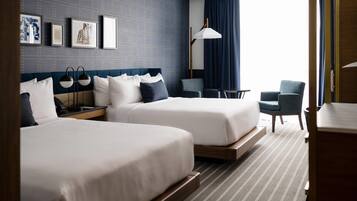 Quarto de hotel com duas camas, uma escrivaninha, uma cadeira, um abajur e vista para o exterior.