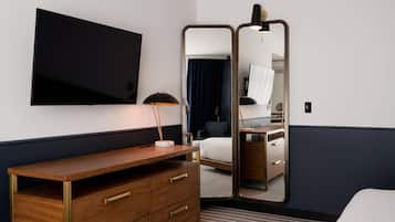 Quarto de hotel com cômoda de madeira, TV de tela plana, escrivaninha com luminária, cama e um espelho grande.