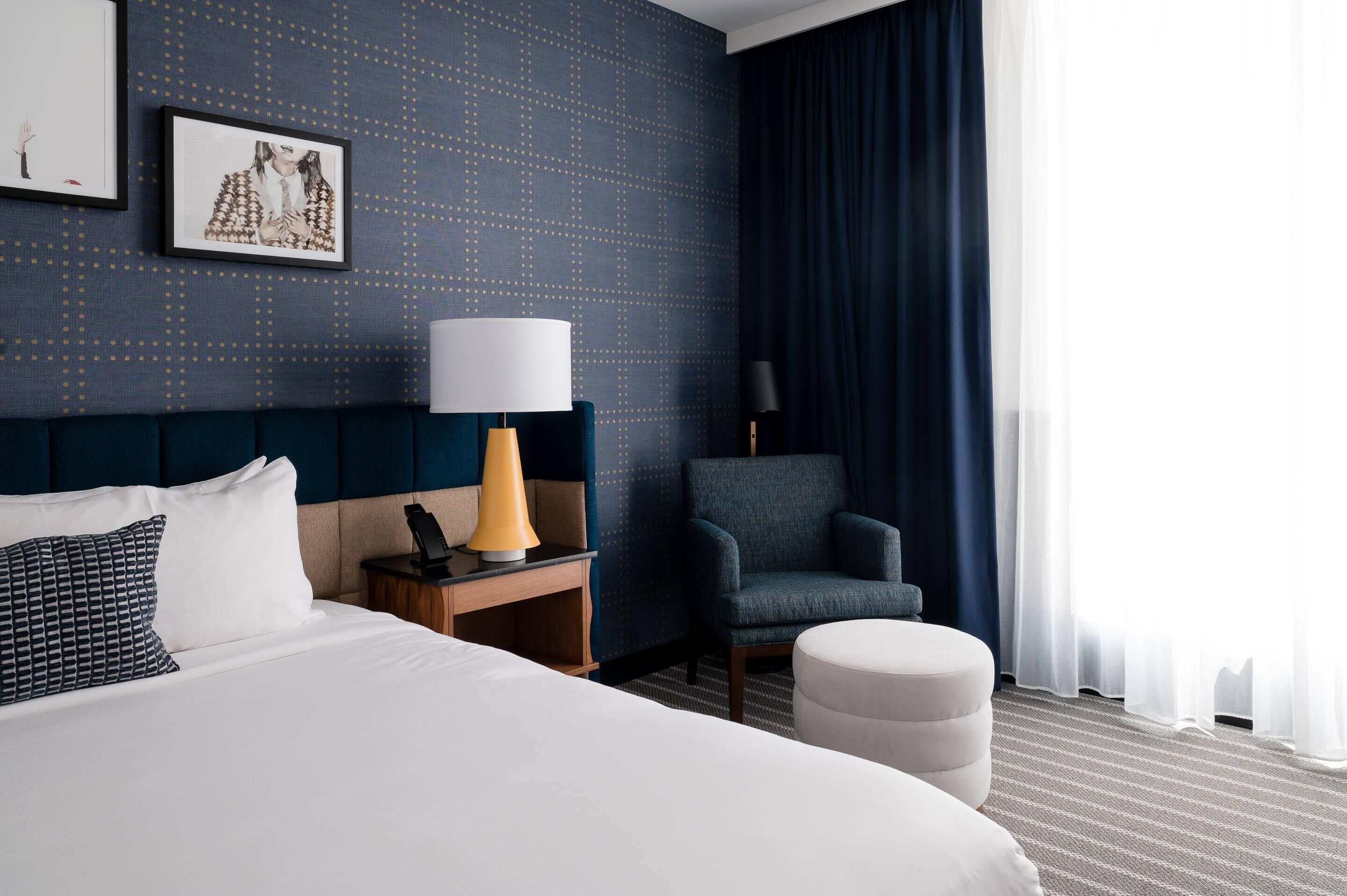 A hotel room with a large bed, a nightstand with a lamp, a chair, and a large window with curtains.