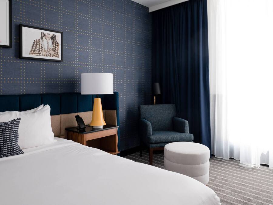 A hotel room with a large bed, a nightstand with a lamp, a chair, and a large window with curtains.