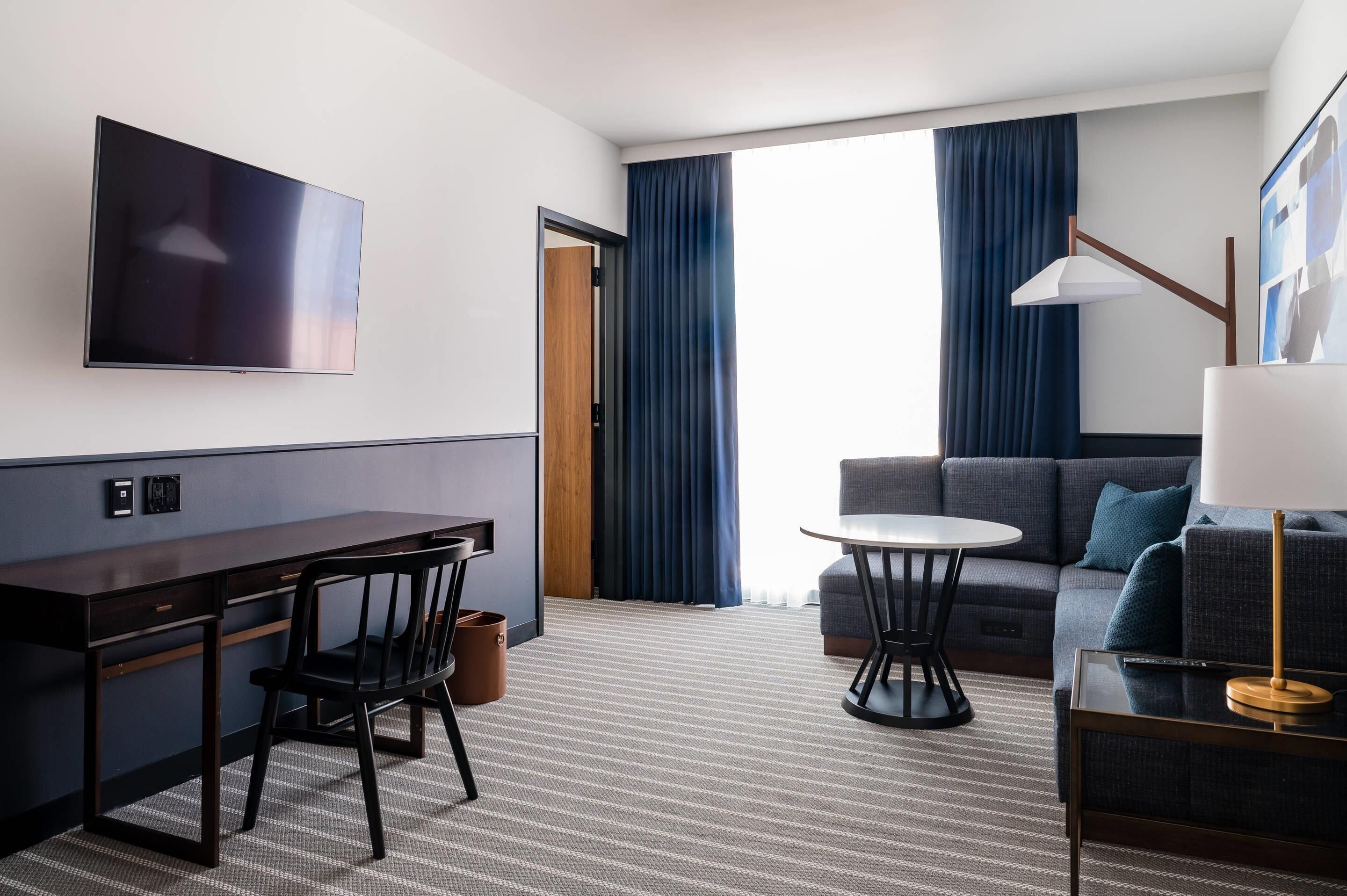 A hotel room with a flat-screen TV mounted on the wall, a desk with a chair, a sofa, a small round table, and a lamp.