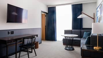 A hotel room with a flat-screen TV mounted on the wall, a desk with a chair, a sofa, a small round table, and a lamp.