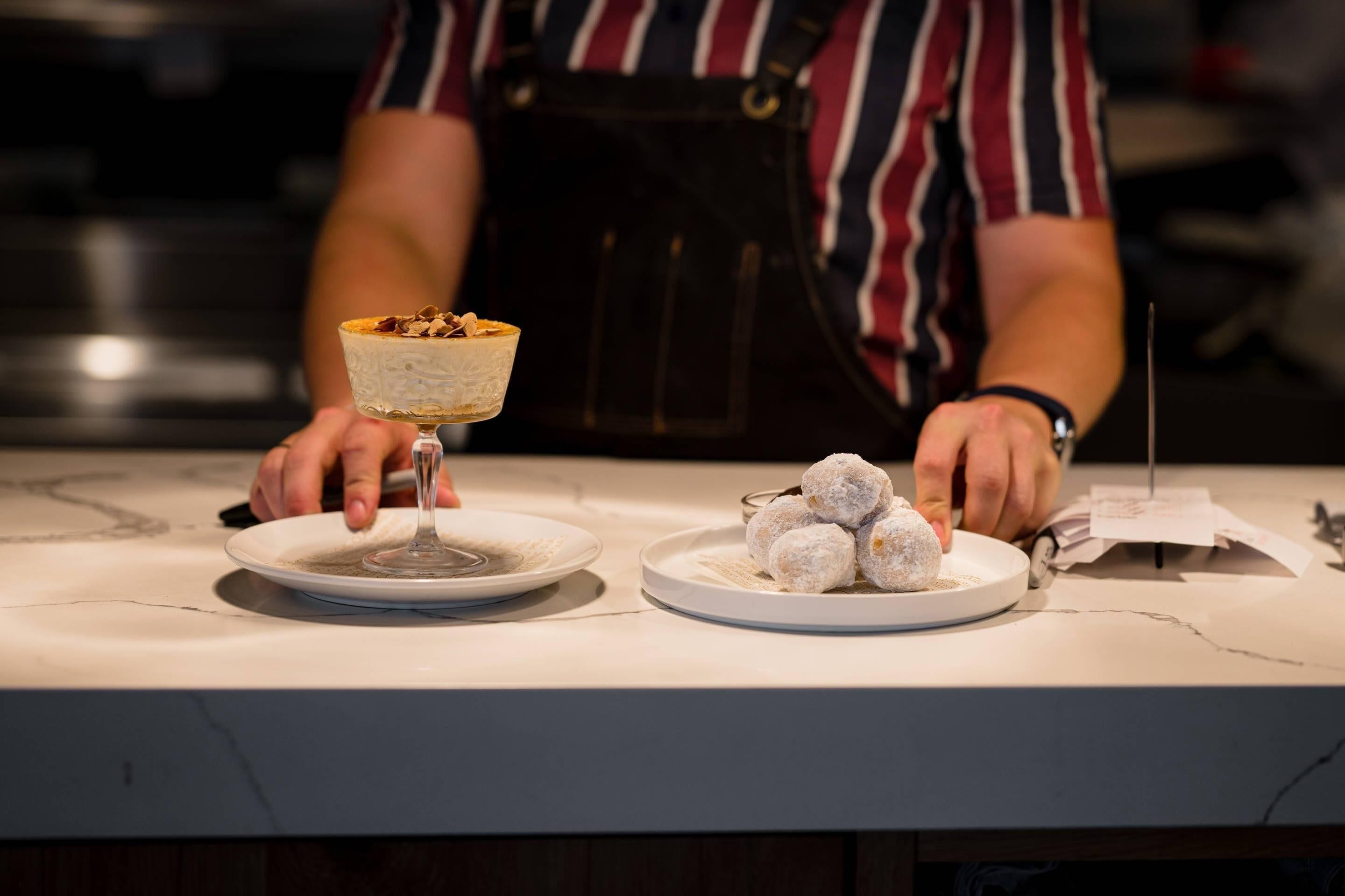 A person holding a dessert in a glass bowl, with a plate of powdered sugar-covered sweets on a marble countertop.