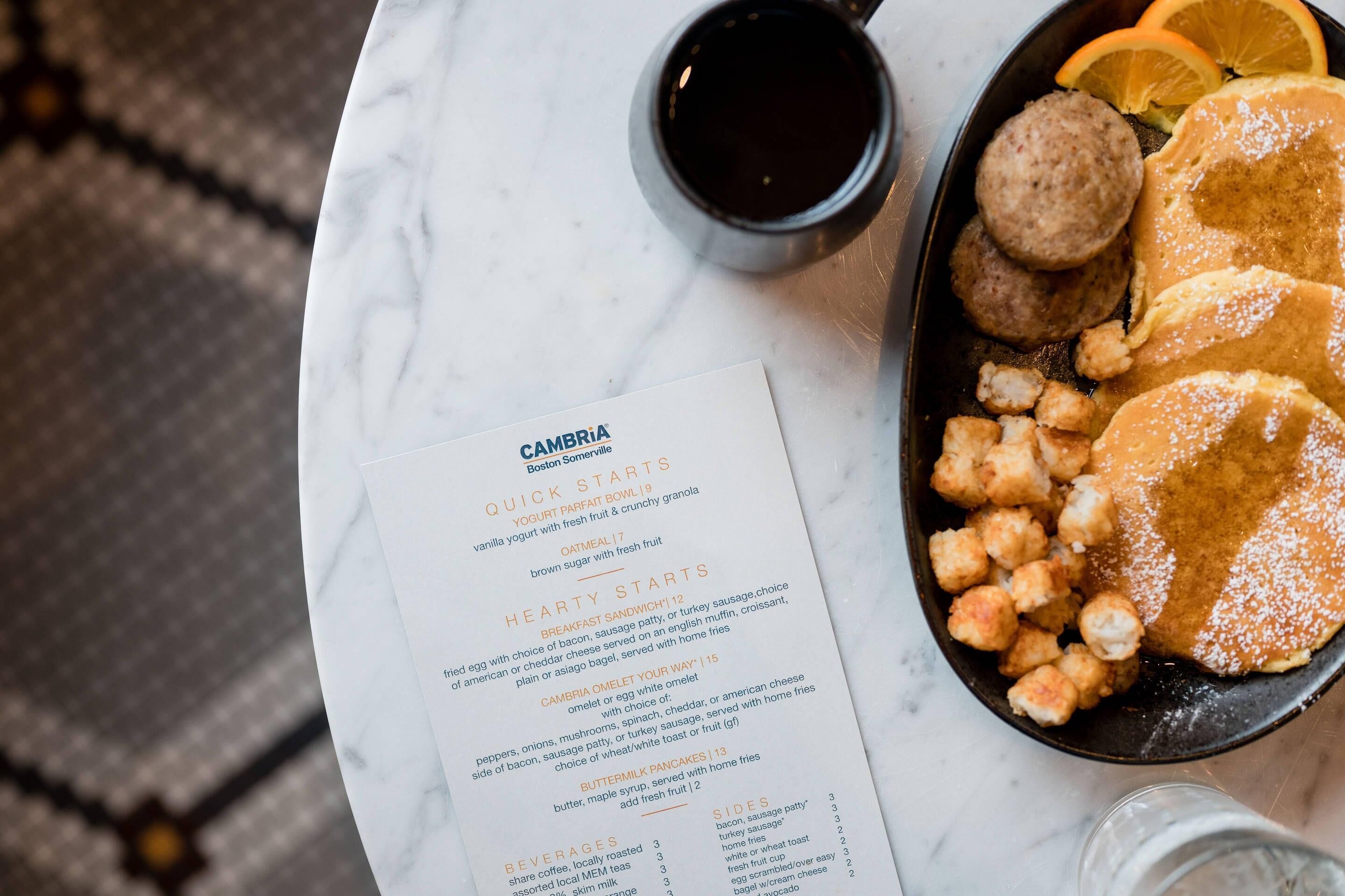 A menu with various breakfast options and sides, a cup of coffee, and a plate of pancakes with orange slices on a marble table.