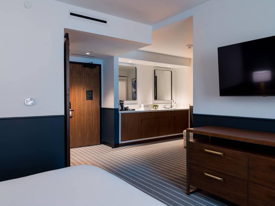 Habitación de hotel con cama, televisor, cómoda de madera, espejo y baño visible a través de una puerta abierta.