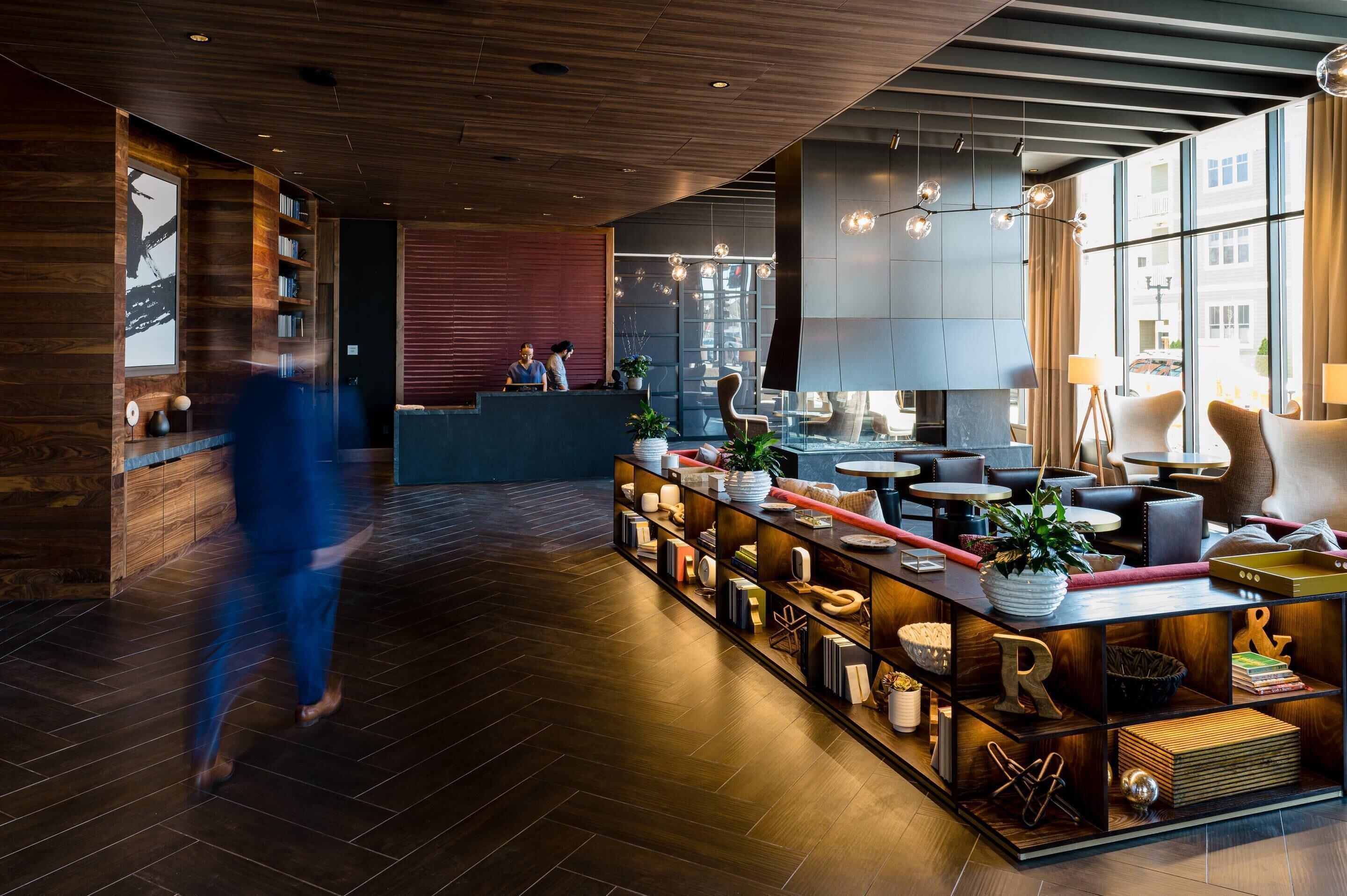 A modern hotel lobby with a wooden floor, a reception desk, and a seating area with tables and chairs.