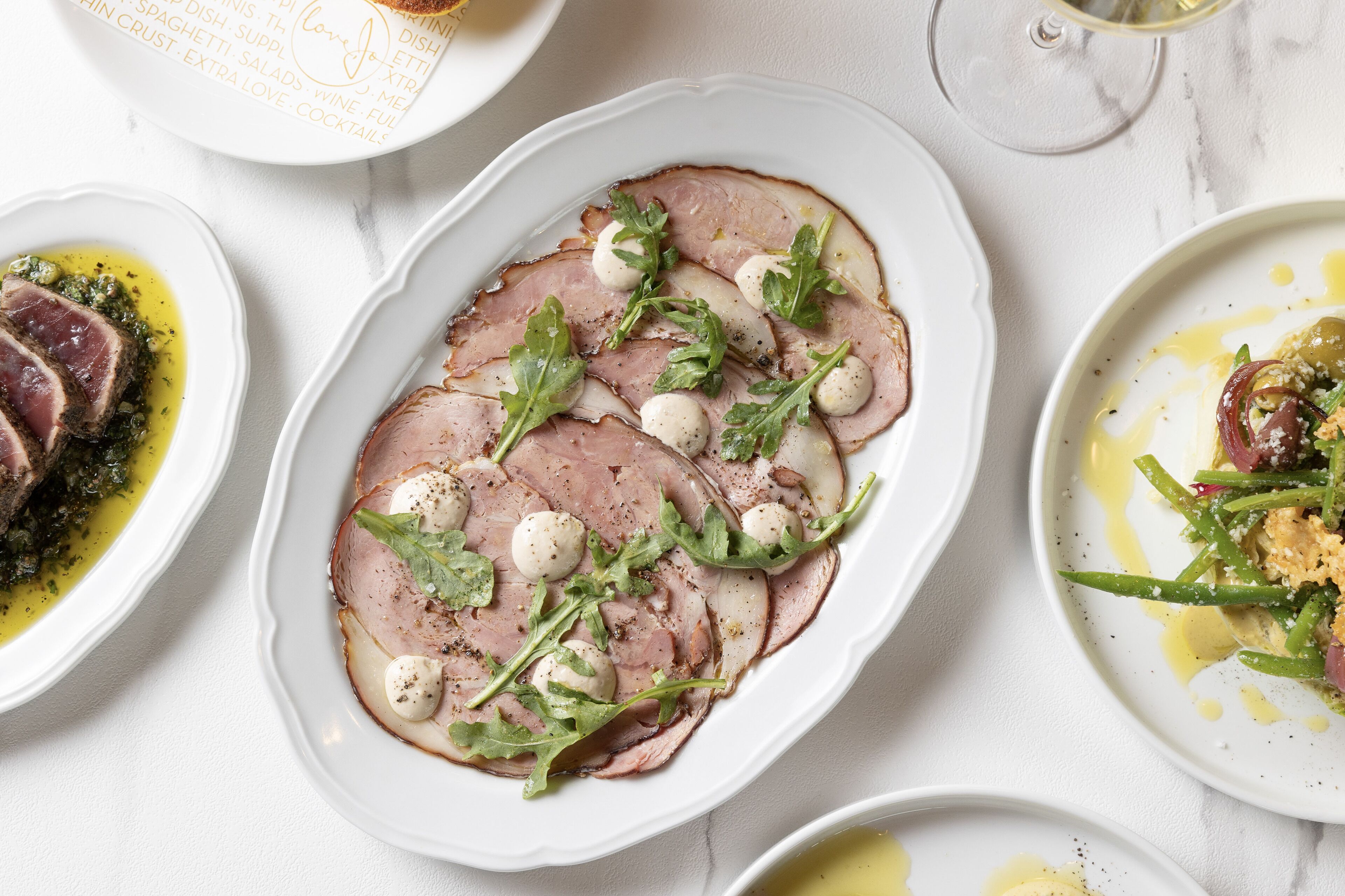 A white table with various dishes, including sliced meat with greens and cheese, and a glass of wine.