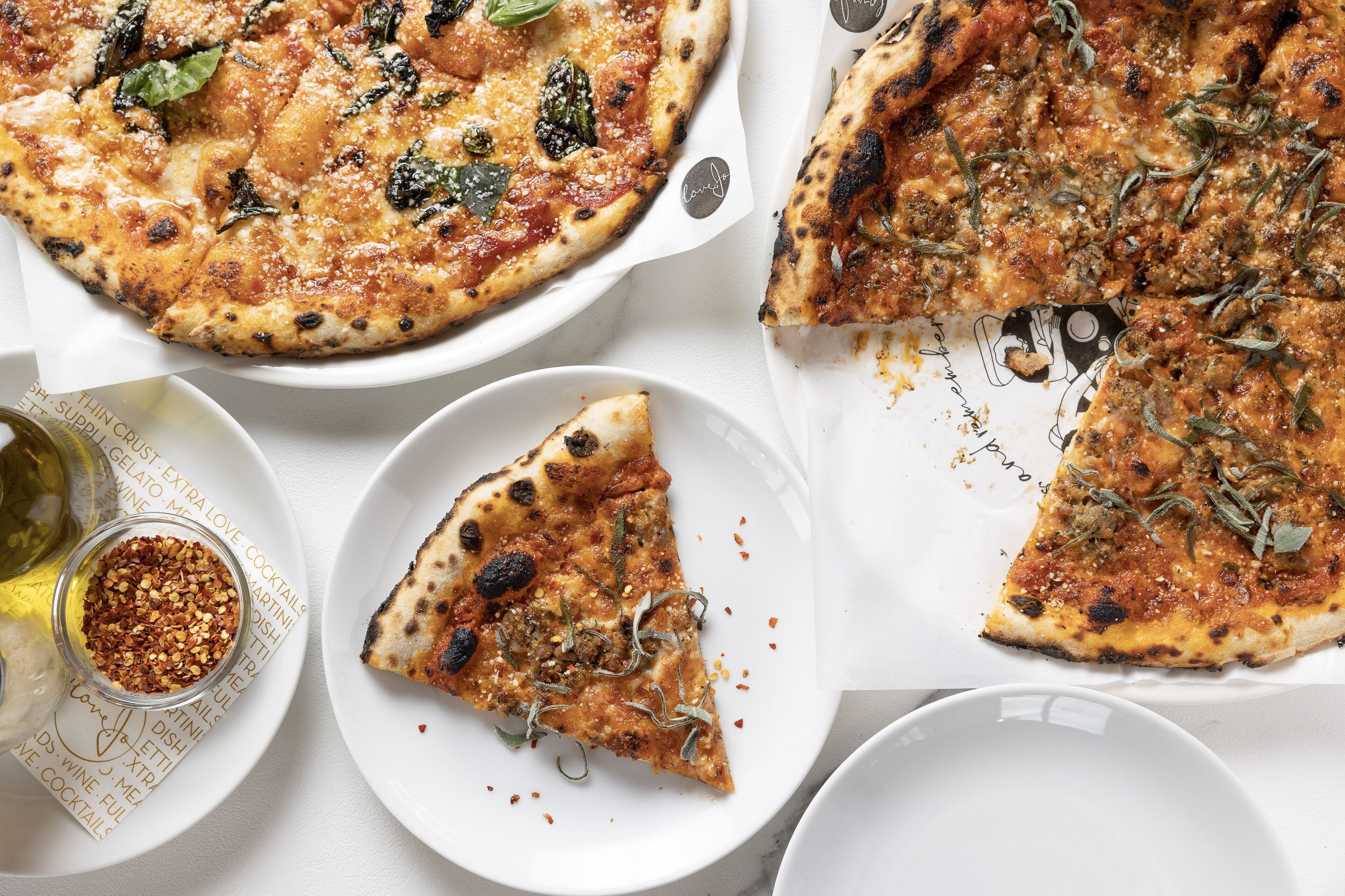 A pizza with various toppings and a slice missing, accompanied by a small bowl of chili flakes and a bottle of oil.
