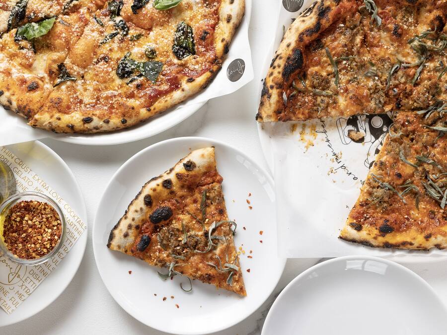 A pizza with various toppings and a slice missing, accompanied by a small bowl of chili flakes and a bottle of oil.