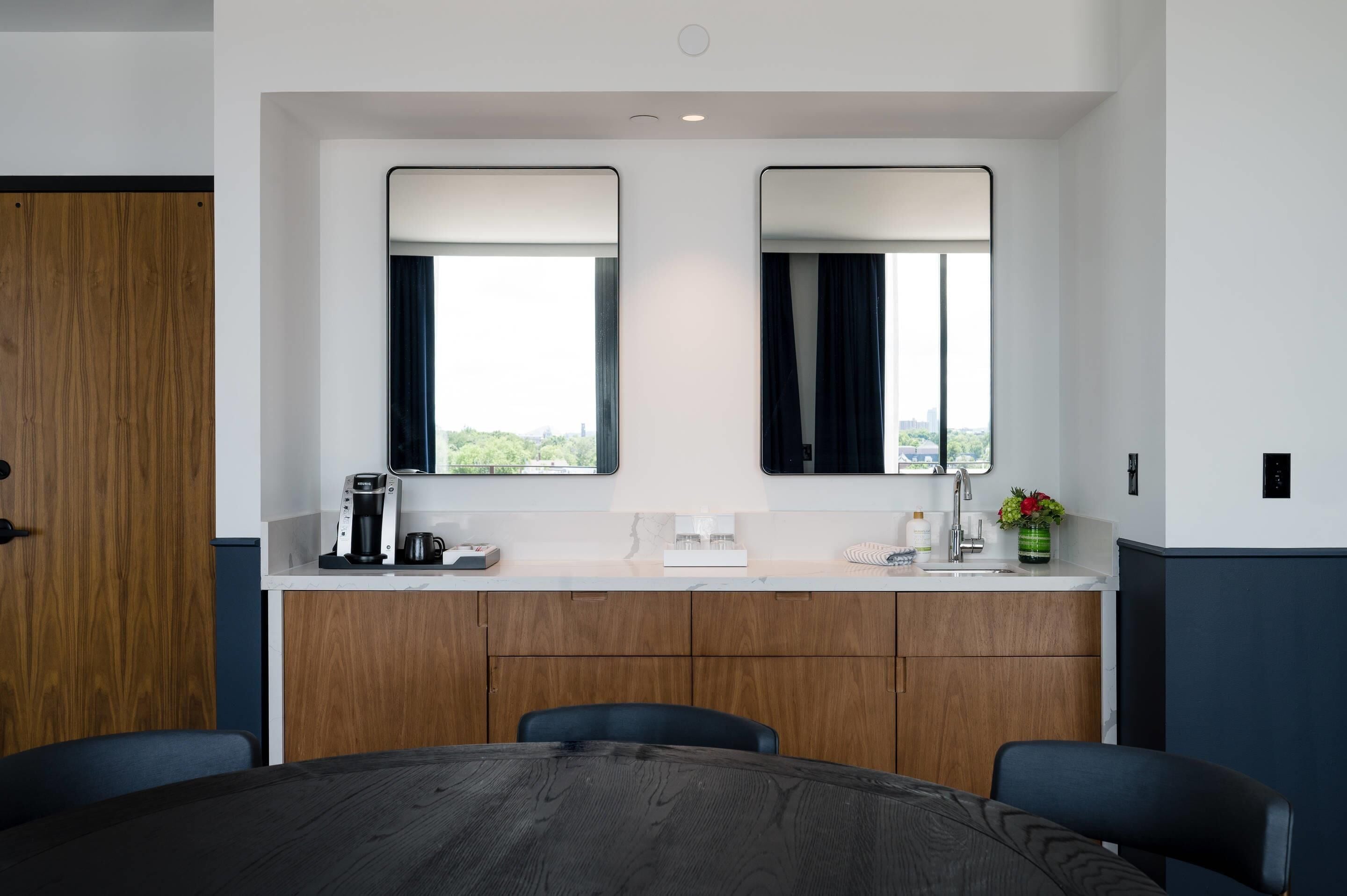 A hotel room with a wooden desk, two mirrors, a coffee maker, a sink, and a view of the city.
