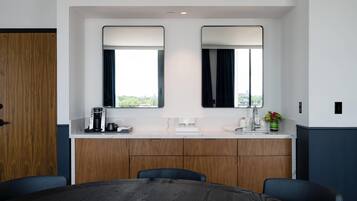 A hotel room with a wooden desk, two mirrors, a coffee maker, a sink, and a view of the city.