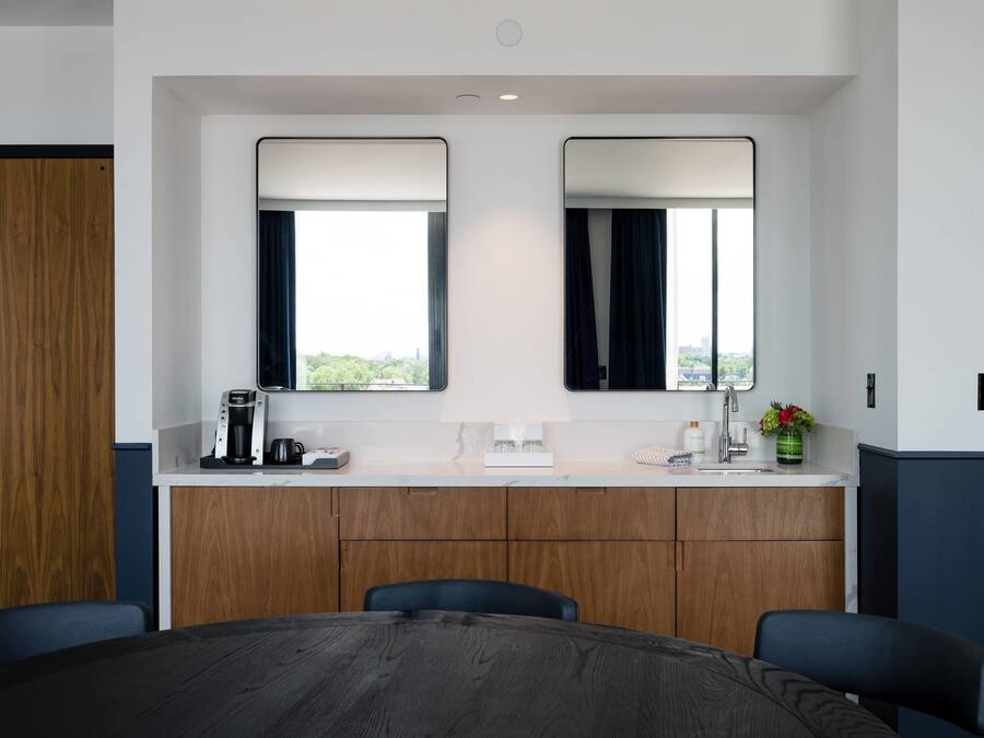 A hotel room with a wooden desk, two mirrors, a coffee maker, a sink, and a view of the city.