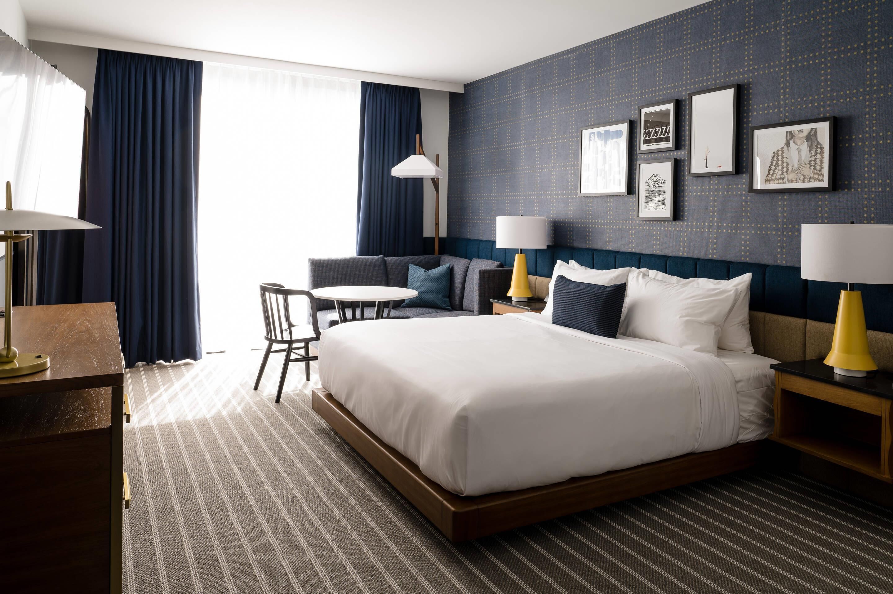 A hotel room with a large bed, a desk, a chair, and a sofa. There are two lamps on the nightstands and a window with curtains.